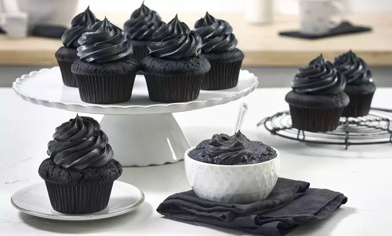 Schokolade-Cupcakes mit glänzender schwarzer Buttercreme auf weißer Tortenplatte, einzelne Cupcakes und Creme in Schale auf schwarzem Tuch drapiert