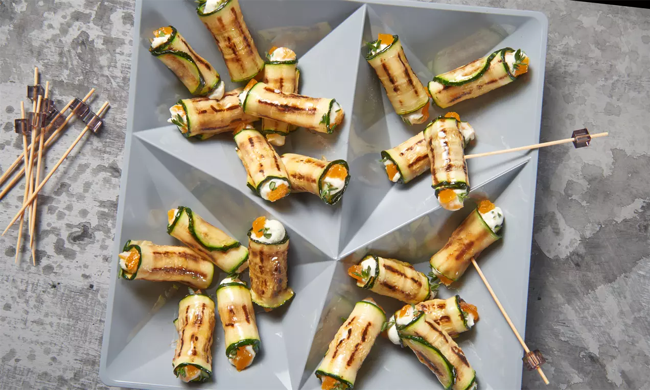 Gerollte Zucchinistreifen mit braunen Grillstreifen, gefüllt mit weißem Frischkäse und oranger Paprika, angerichtet auf einem grau-eckigen Teller