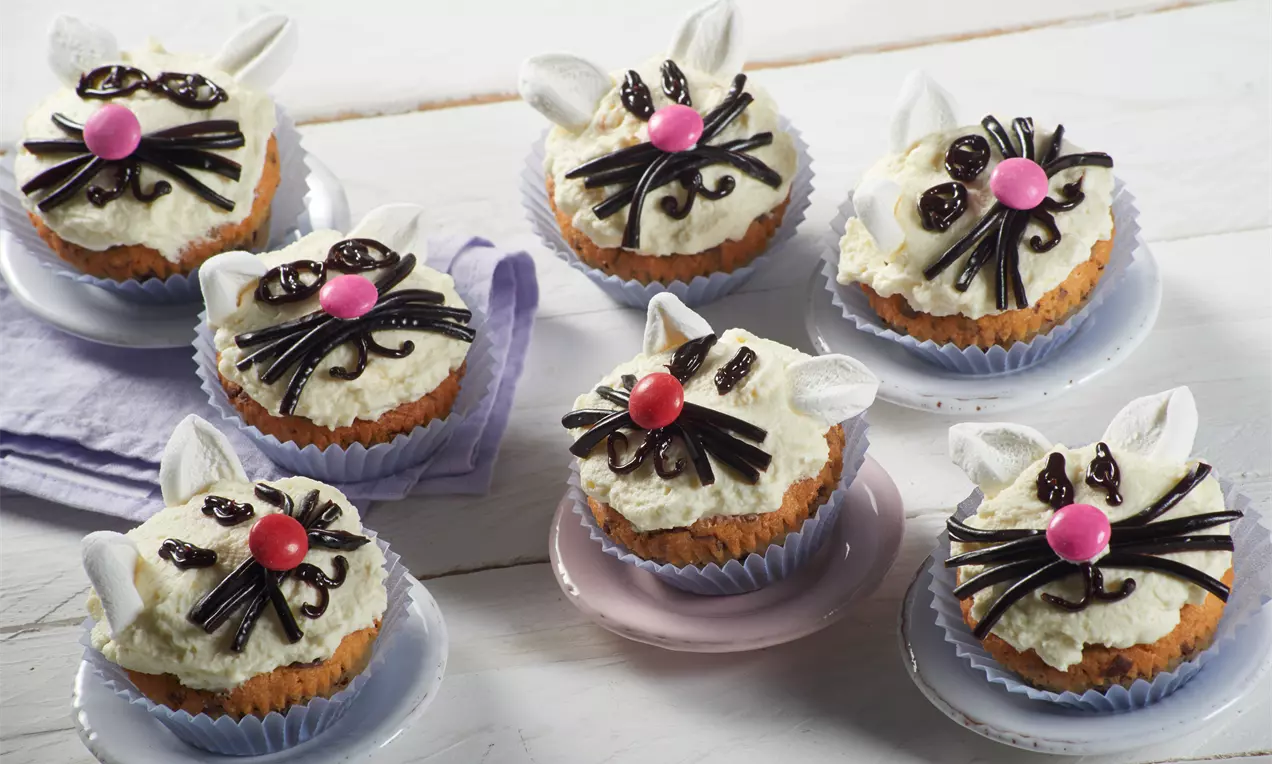 Muffins mit weißer Creme, Fondant-Ohren, schwarzen Lakritz-Streifen als Schnurrhaare und rosa Zuckerperlen auf bunten Tellerchen