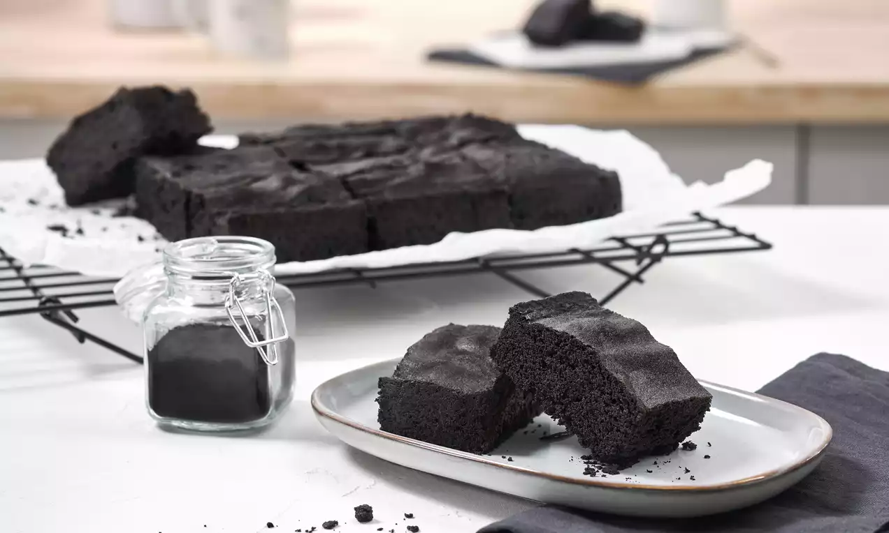 Zwei dunkle, saftige Brownie-Stücke mit glatter Oberfläche auf einem weißen Teller neben einem Glas mit schwarzem Pulver