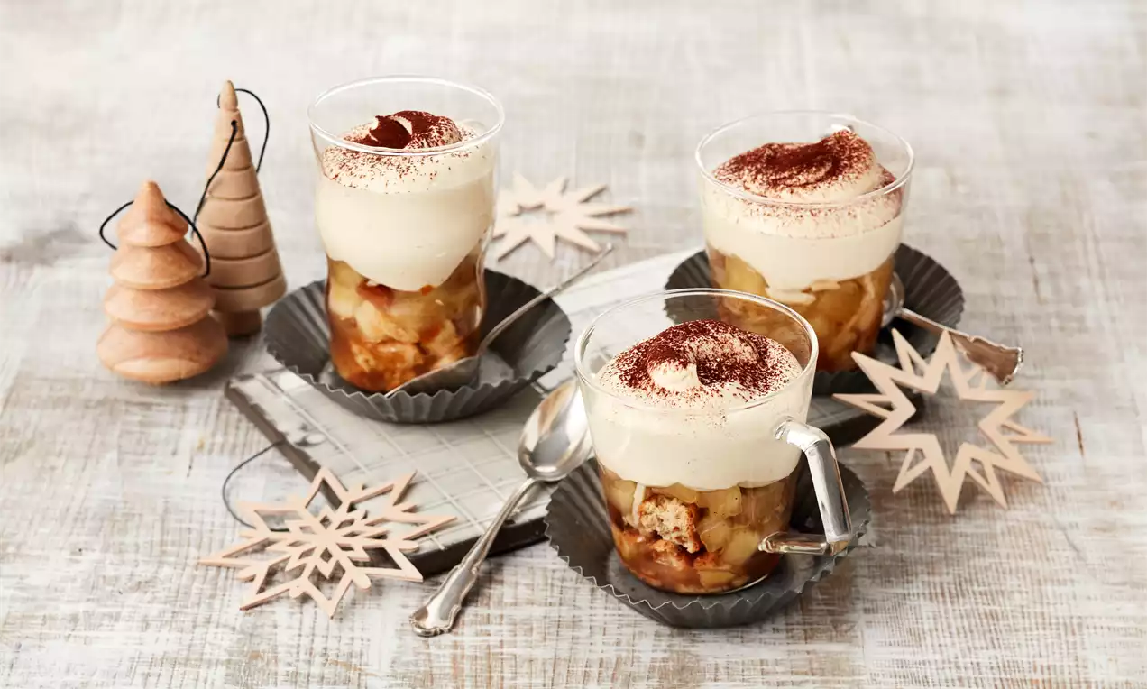 Bratapfel-Tiramisu mit cremiger Schicht, Apfelstücken und Biskuit in Gläsern auf dunklen Tellern, dekoriert mit Sternen und Holztannen