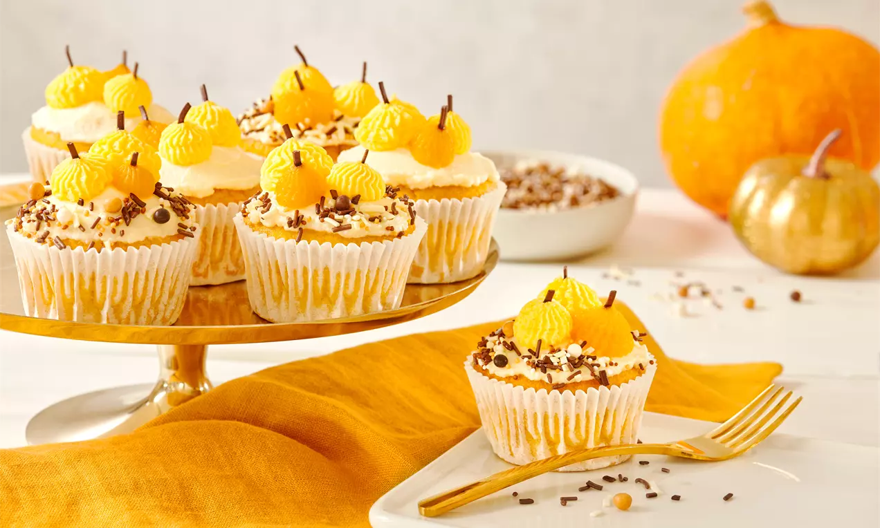 Kürbis-Cupcakes mit weißer Cremehaube, orangenen Marzipankugeln, Schokostreuseln und Herbstdeko auf goldener Etagere und Teller arrangiert