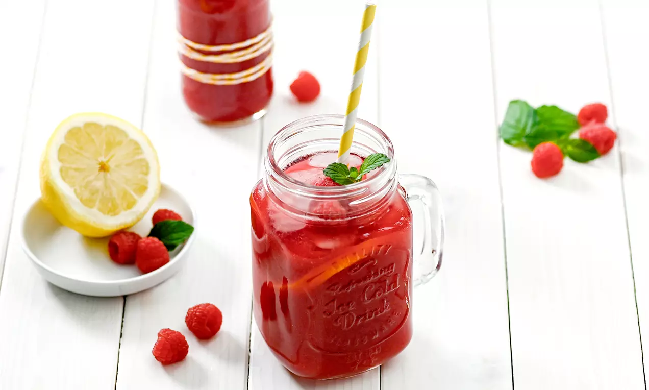 Himbeerrote Limonade mit Eiswürfeln und Minzblättern in einem Glas mit Gelb-Weiß-Strohhalm neben frischen Himbeeren und Zitronenscheibe