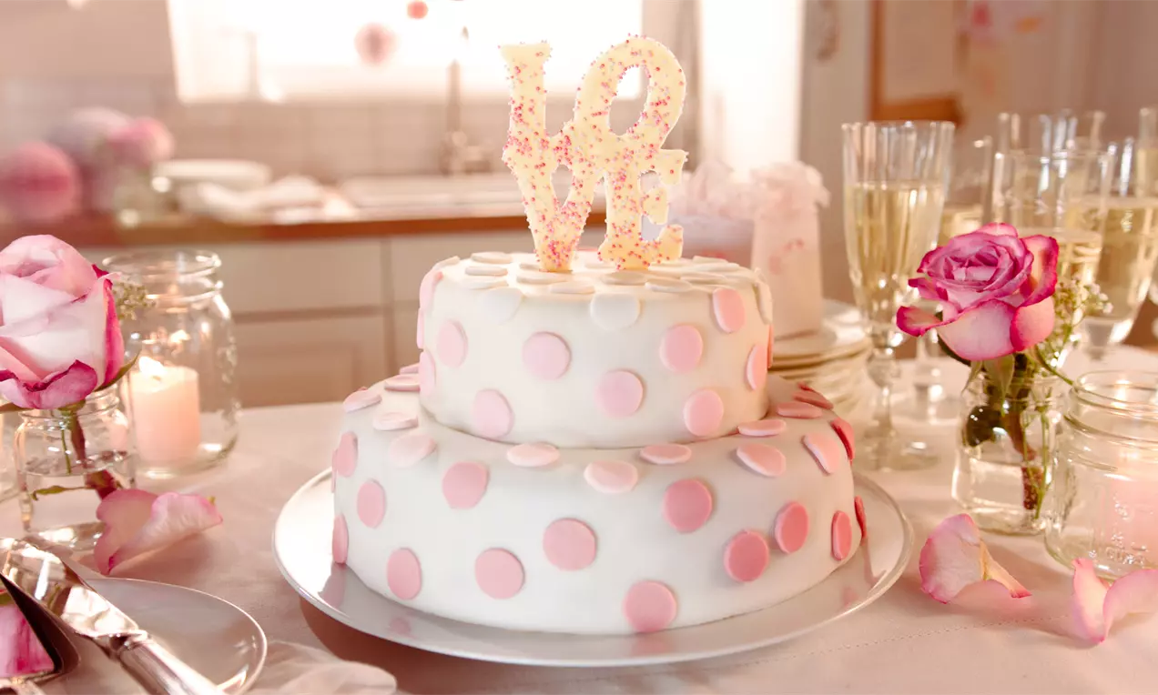Zweistöckige Torte mit weißem Fondant und rosafarbenen Punkten, gekrönt von LOVE-Dekor, auf einer silbernen Platte mit Rosen und Gläsern dekoriert