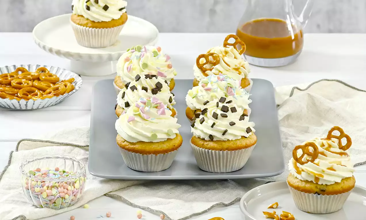 Cupcakes in Papierförmchen mit goldbraunem Boden und lockerem Sahne-Topping, garniert mit bunten Streuseln, Schokostückchen und Brezeln auf grauem Teller