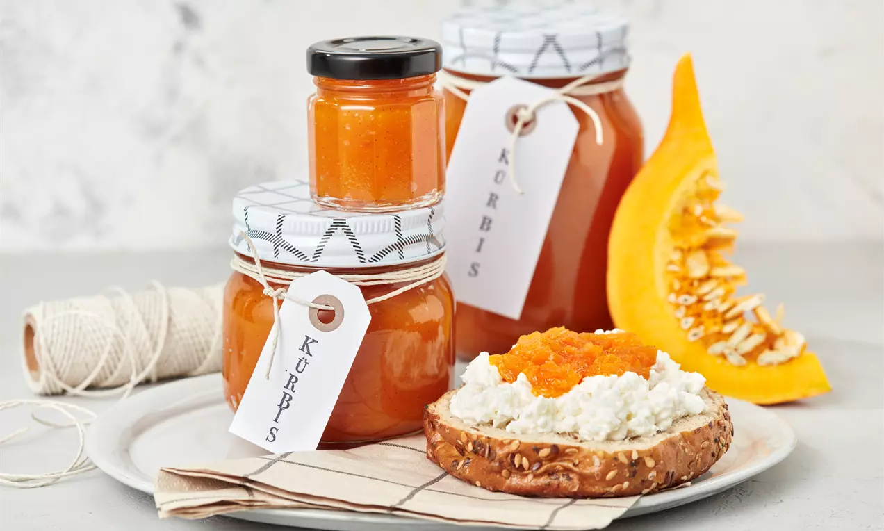 Gläser mit orangefarbener Kürbismarmelade auf einem Teller, dekoriert mit Kürbisscheibe, Brot mit Frischkäse und Marmelade sowie Kordel