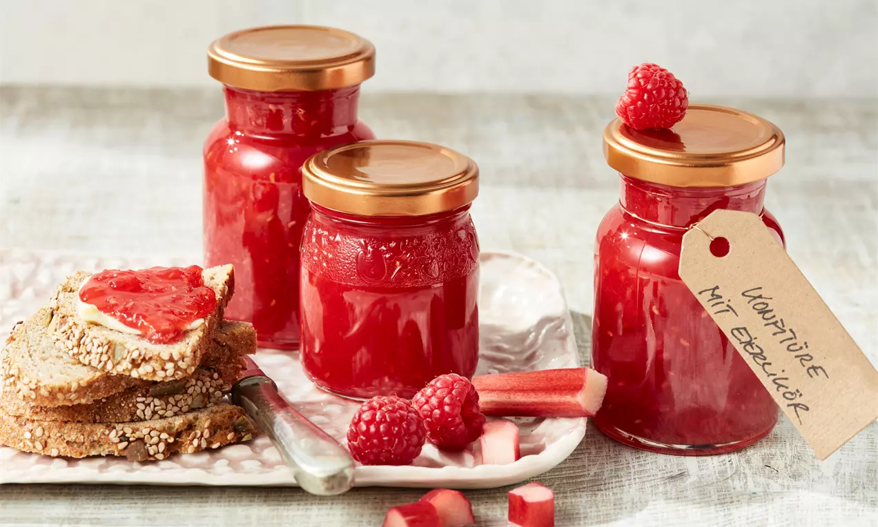 Drei gläser rote Marmelade mit goldenen Deckeln, dekoriert mit Himbeeren und Rhabarberstangen, daneben Vollkornbrot mit Marmelade bestrichen