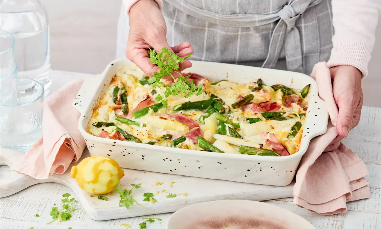 Goldbraune Spargellasagne mit grünem und weißem Spargel, Schinken und geschmolzenem Käse in weißer Keramikform, von frischer Kresse gekrönt
