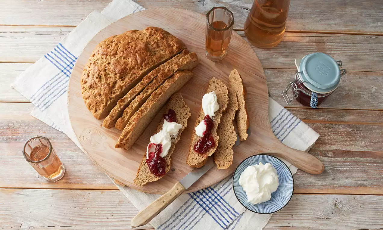 Goldbraunes, rustikales Anisbrot auf einem runden Holzbrett, mit Scheiben serviert, die mit Sahne und roter Marmelade garniert sind