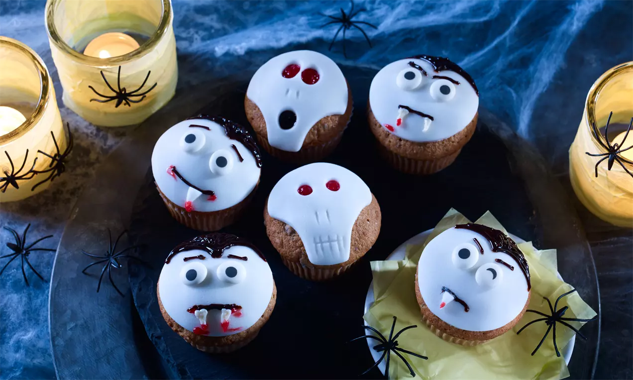 Sechs Muffins mit weißem Zuckerguss als Totenköpfe und Vampire verziert, auf schwarzem Teller mit Spinnennetzen und Kerzen drapiert