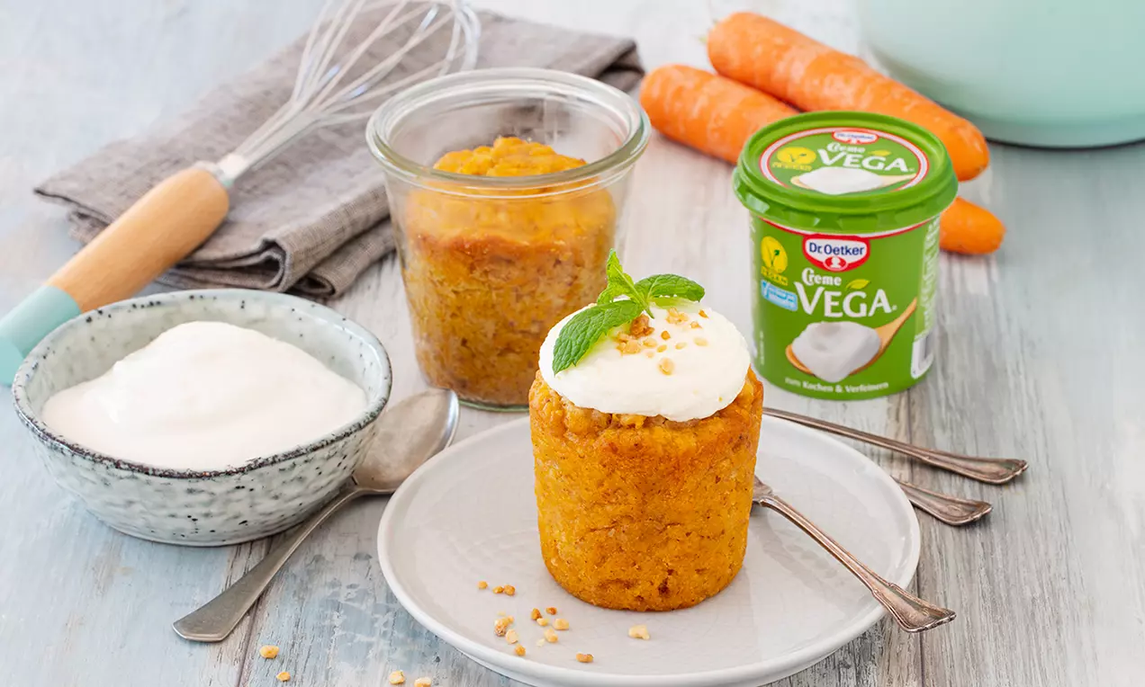 Leuchtend orangefarbener Möhrenkuchen in einem Glas und auf einem weißen Teller, dekoriert mit weißem Frosting, Minzblatt und gehackten Nüssen