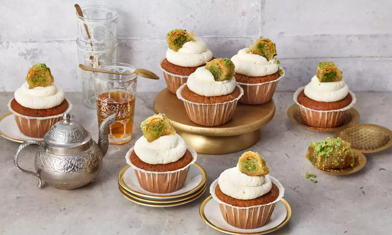 Baklava-Cupcakes mit goldbraunem Teig, weißer Sahnehaube und einem grünen Pistazien-Baklava-Stück auf Tellern und einer goldenen Platte arrangiert