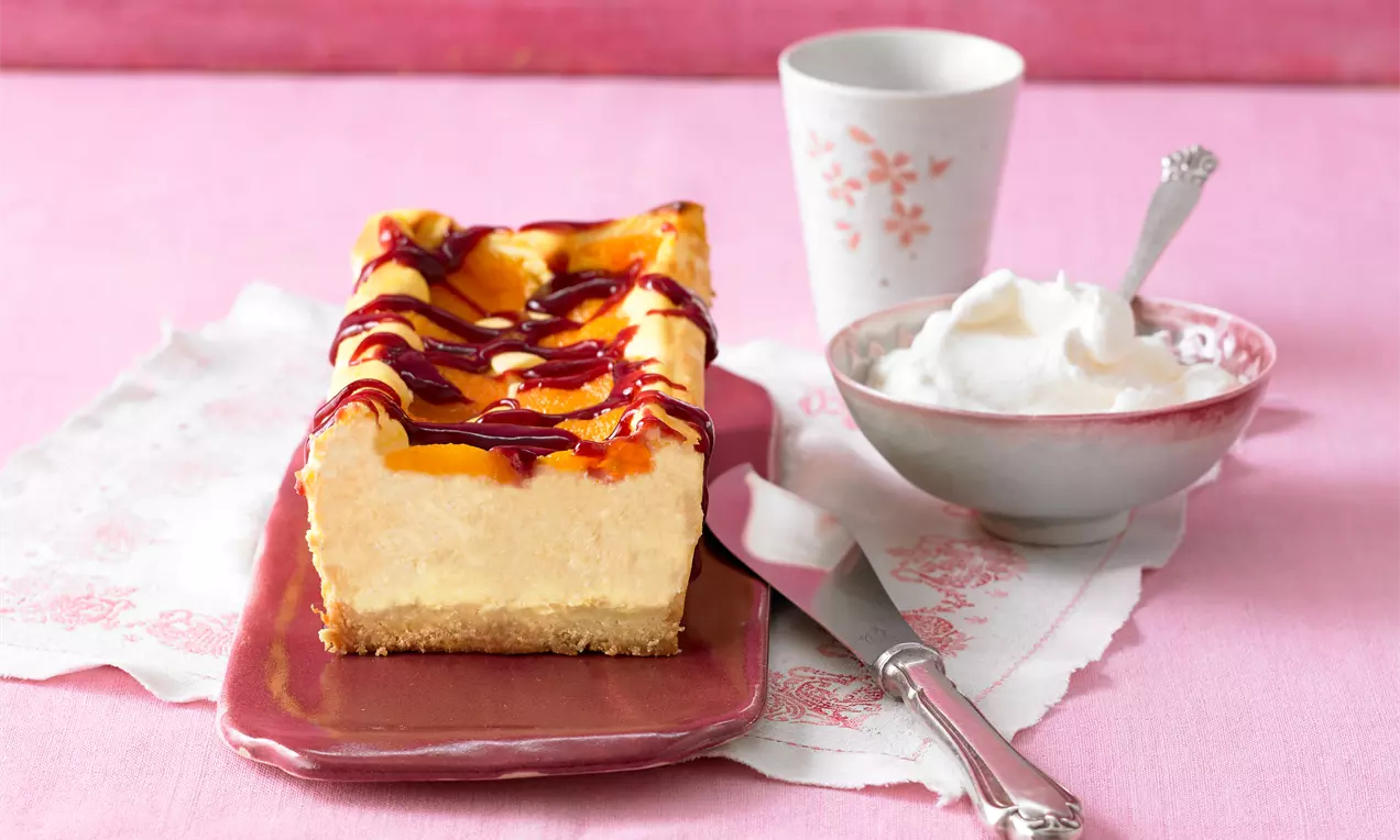 Aprikosen-Käsekuchen mit Himbeersprenkeln auf weißer Platte, cremige Füllung, goldene Aprikosenstücke und rote Himbeerspritzer