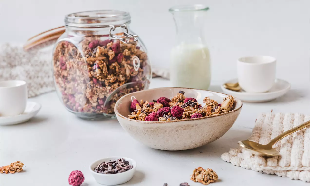 Knuspriges Granola mit goldbraunen Clusterstücken und roten Beeren, angerichtet in einer beige-matten Schale auf hellem Untergrund, AI-labeled image