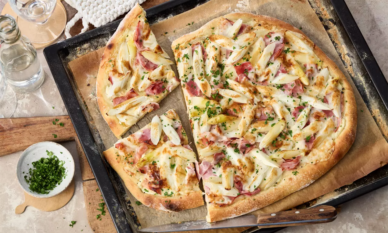 Goldbraune Spargelpizza mit knusprigem Rand, belegt mit hellem Spargel, Schinkenstreifen, Käse und frischen Kräutern auf Backpapier