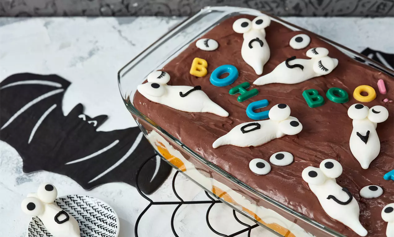 Halloween-Dessert in einer Glasform mit dunkelbrauner Creme, Geisterfiguren aus weißem Dekor und bunten Buchstaben auf schwarzem Stoff