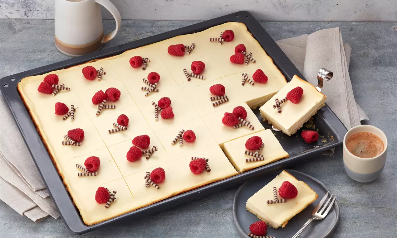 Käsekuchen vom Blech, hellgoldene Cremefüllung in Quadrate geschnitten, mit roten Himbeeren und feinen Schokoröllchen dekoriert auf grauem Untergrund
