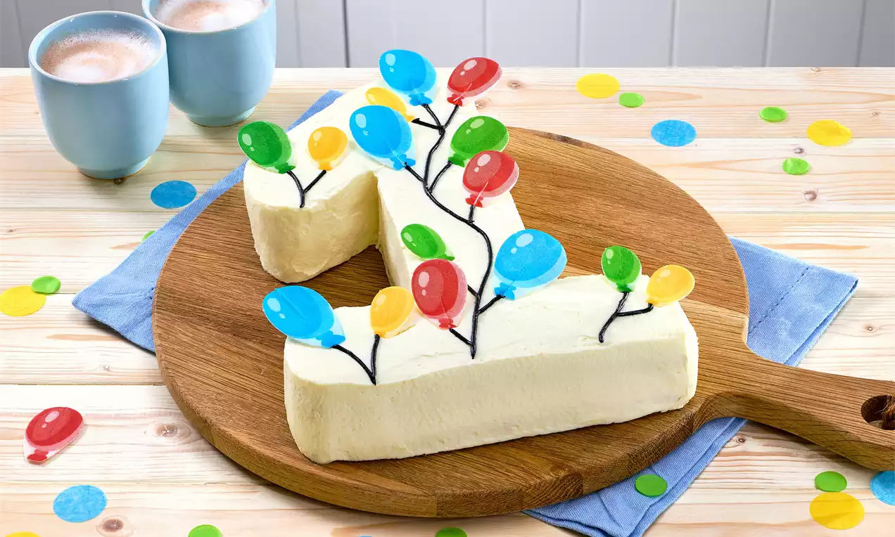 Gebogener Geburtstagskuchen in Zahl-Form, mit heller Creme überzogen, bunten Luftballon-Dekoren und Holzplatte auf hellem Holztisch