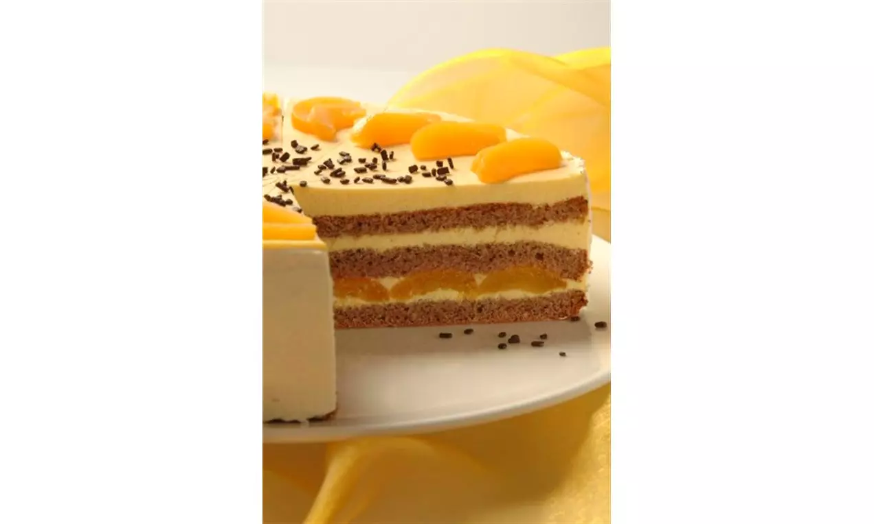 Aprikosensekttorte mit gelben Obststücken und Schokostreuseln auf heller Creme, geschichtet zwischen braunen Kuchenteigen auf weißer Platte