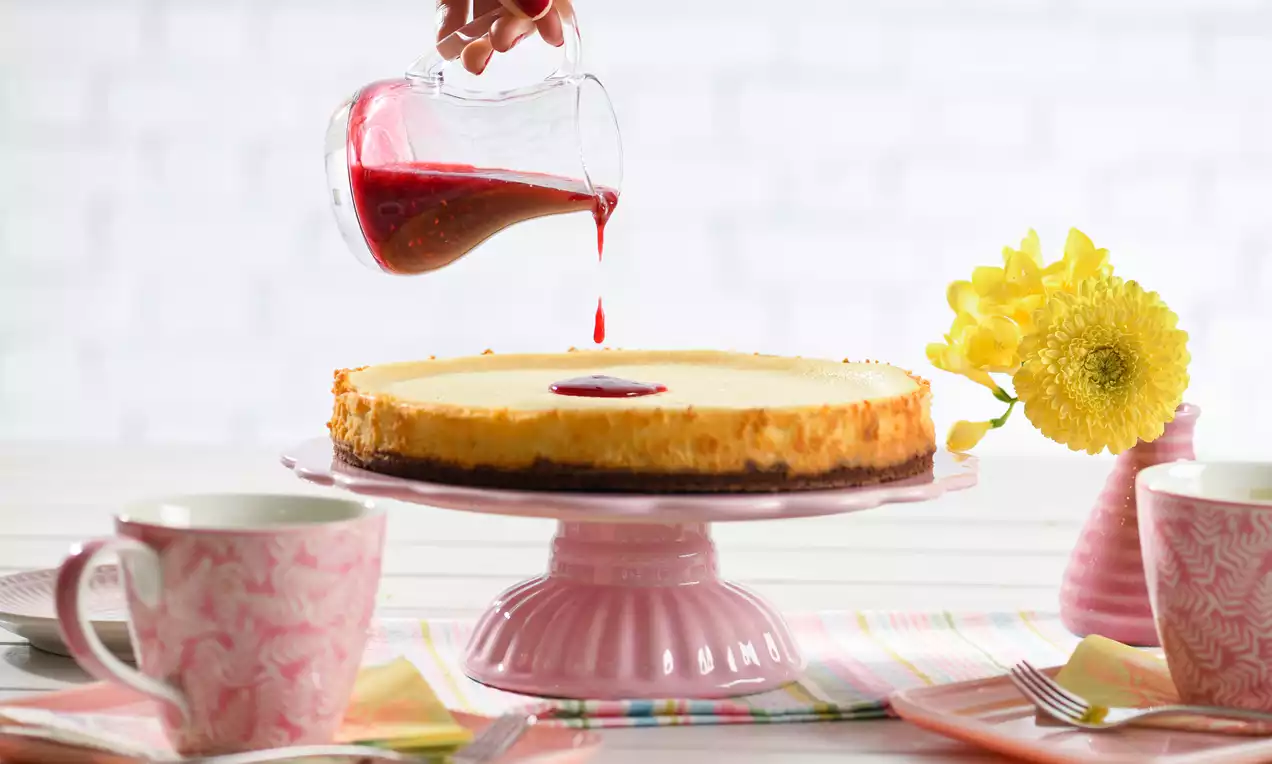 White-Chocolate-Cheesecake auf rosa Tortenständer mit goldgelber Oberfläche und dunklem Boden, garniert mit roter Sauce, daneben gelbe Blumen