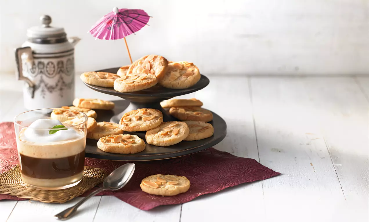 Goldbraune Mandelplätzchen mit gesplitterter Oberfläche auf schwarzer Etagere, daneben Kaffee im Glas und Dekoration mit rosa Schirmchen