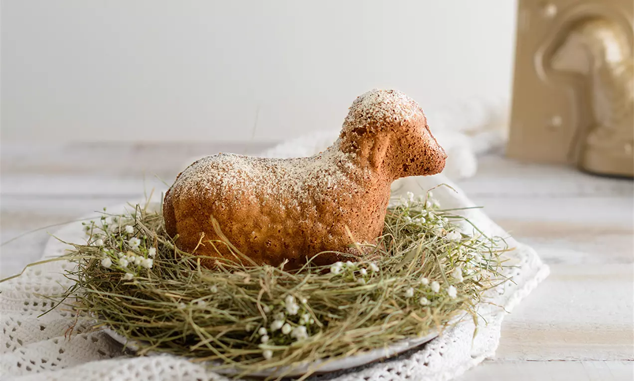 Goldbraunes Osterlamm aus Biskuitteig, mit Puderzucker bestäubt, ruhend auf einem Nest aus Heu mit weißen Blüten auf einem Teller