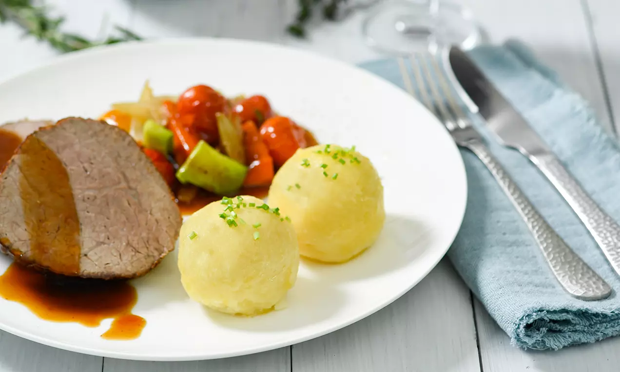 Zwei goldgelbe Kartoffelknödel mit fein gehacktem Schnittlauch bestreut, serviert neben Bratensaft, Gemüse und Fleischscheibe auf weißem Teller