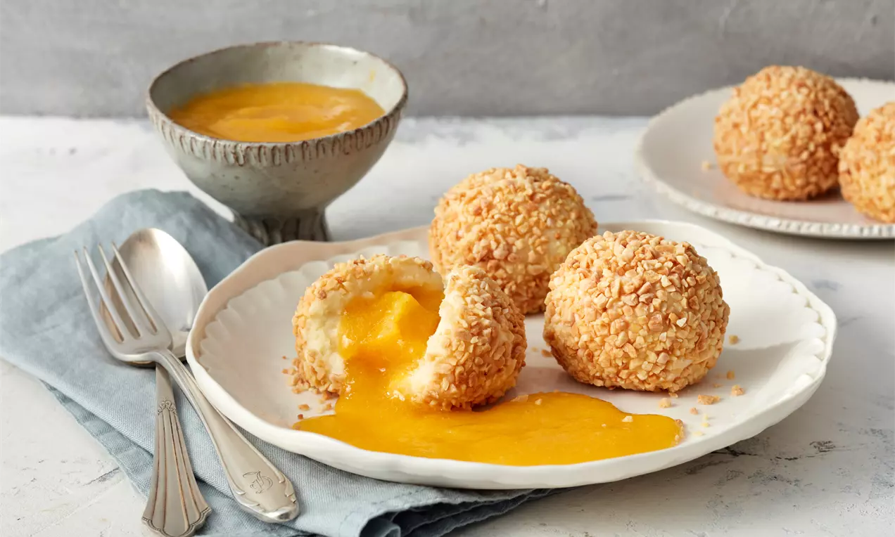 Goldgelbe, mit gehackten Nüssen bestreute Grießknödel auf weißem Teller, einer halb geöffnet mit fließender orangefarbener Füllung.