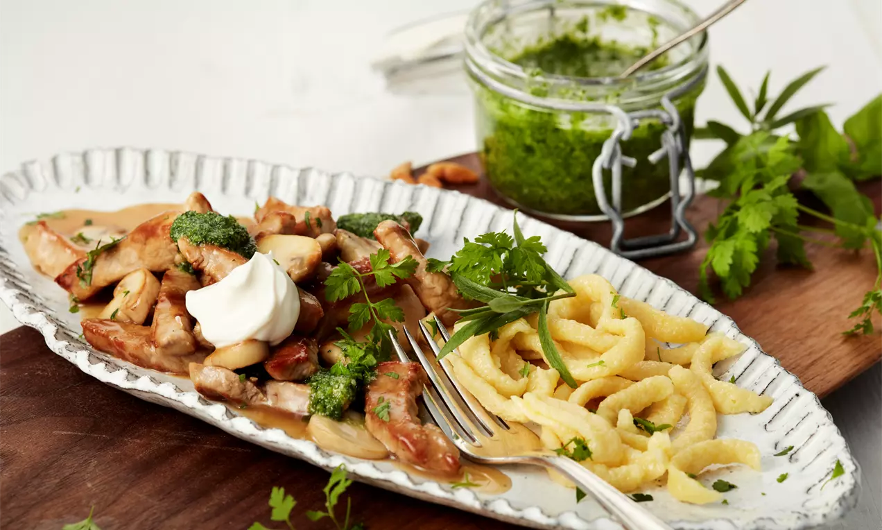 Gebratenes Geschnetzeltes mit grünem Pesto und Sahnehäubchen neben goldgelben Spätzle, serviert auf einem weißen Teller mit frischen Kräutern