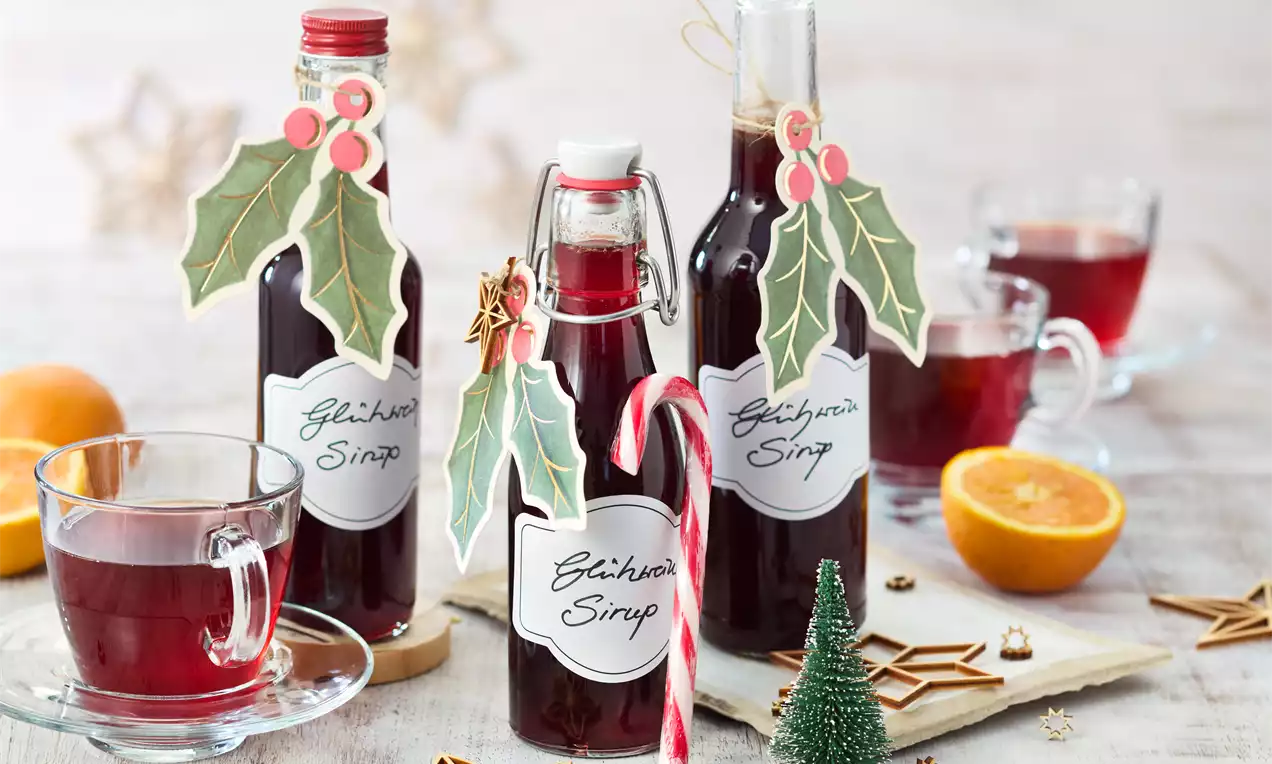 Dunkelroter Glühweinsirup in Flaschen mit weihnachtlicher Deko aus Ilex, umgeben von Glühweingläsern, Orangenscheiben und Holzsternen