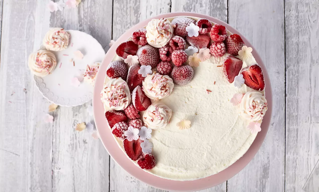 Eistorte mit weißer glatter Oberfläche, dekoriert mit roten Beeren, rosa Baisertupfen und kleinen Blüten auf rosa Teller