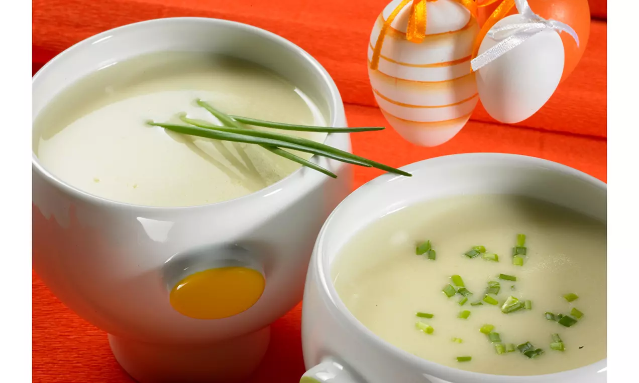 Parmesansuppe mit cremiger hellgelber Konsistenz, Kartoffelstücken, geriebenem Parmesan, Schnittlauch und Croutons in weißer Schale