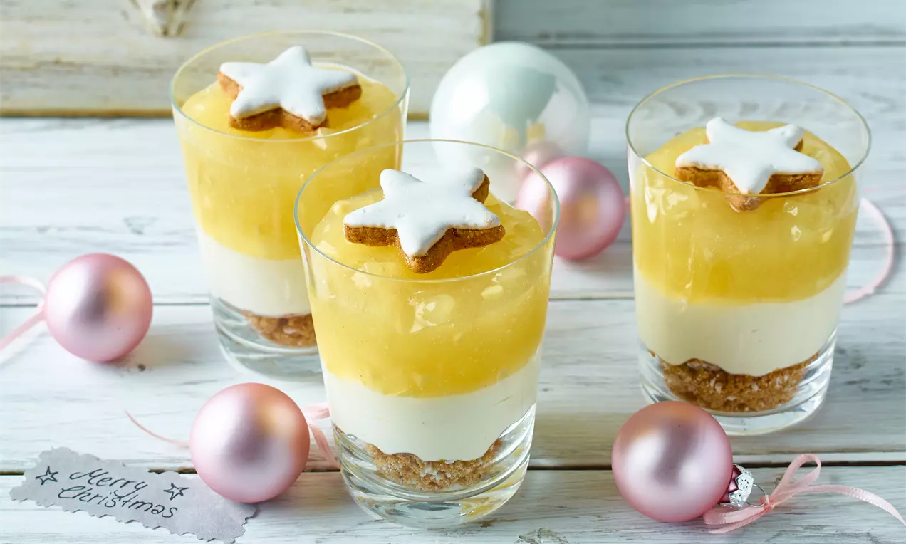 Weihnachtliches Quarkdessert mit cremiger weißer Schicht, gelber Wintergrütze, Zimtstern-Topping in Gläsern, umgeben von rosa Weihnachtskugeln