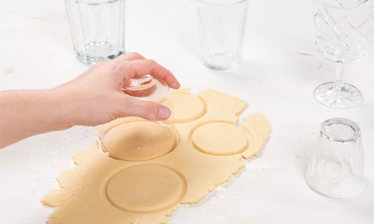 Hand, die mit einem Glas Kreise aus einem hellen, glatten Teig aussticht, auf einer weißen Arbeitsfläche mit verstreuten Gläsern