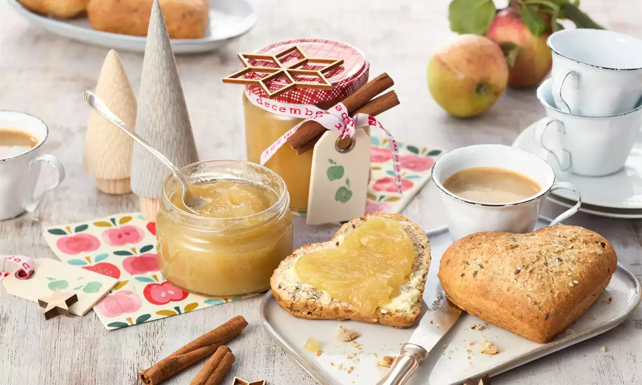 Apfel-Zimt-Marmelade in einem offenen Glas und dekorativem Schraubglas mit rot-weißem Deckelband, daneben Brötchenherz mit Marmelade bestrichen
