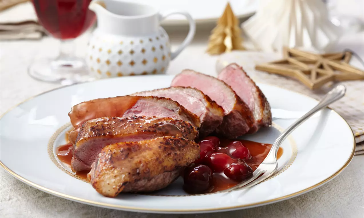 Entenbrust mit knuspriger, goldbrauner Haut und zarten rosa Scheiben auf Orangen-Cranberry-Soße, garniert mit roten Cranberries auf weißem Teller