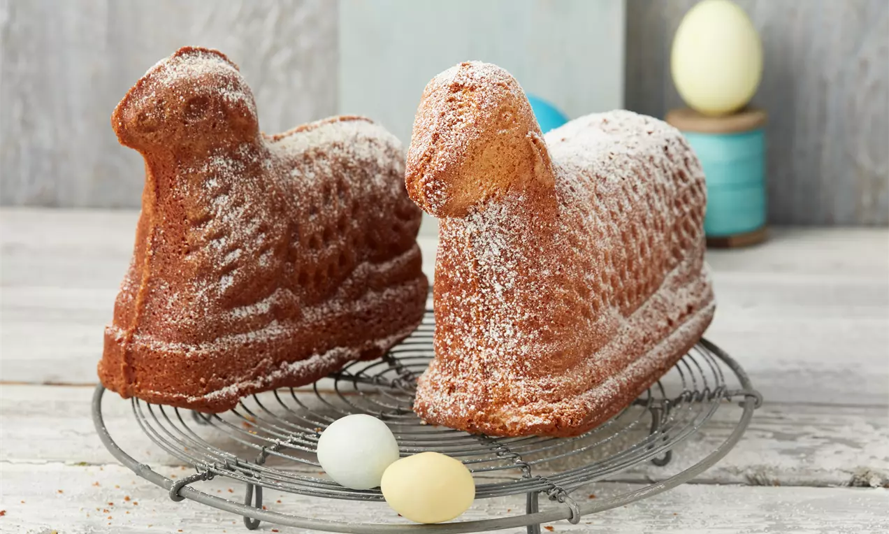 Zwei goldbraune Osterlämmer aus Rührteig mit feiner Puderzuckerhaube stehen auf einem runden Kuchengitter neben pastellfarbenen Deko-Eiern