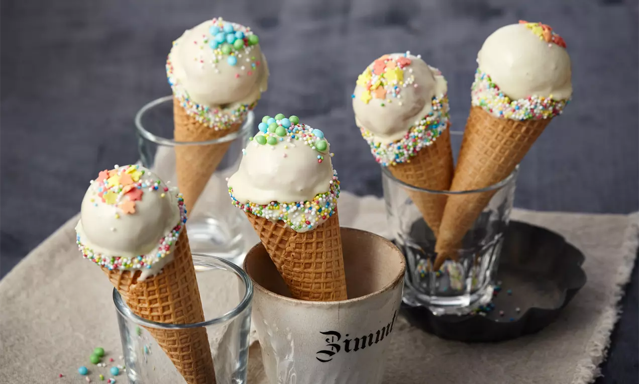 Cake-Pops in Waffelhörnchen mit heller Glasur und bunten Streuseln, aufrecht in Gläsern und einem Papierförmchen auf Stoff drapiert