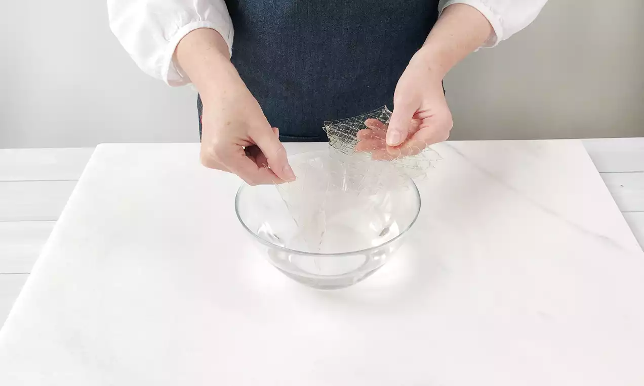 Eine Person steckt durchsichtige, wellige Gelatineblätter in eine klare Glasschale mit Wasser auf einer weißen, marmorierten Oberfläche