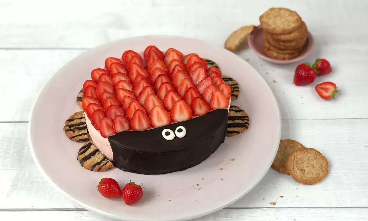 Frischkäse-Erdbeer-Torte mit leuchtend roten Erdbeerscheiben, dunkler Schokoladenglasur und Augen, umgeben von Keksen auf weißem Teller