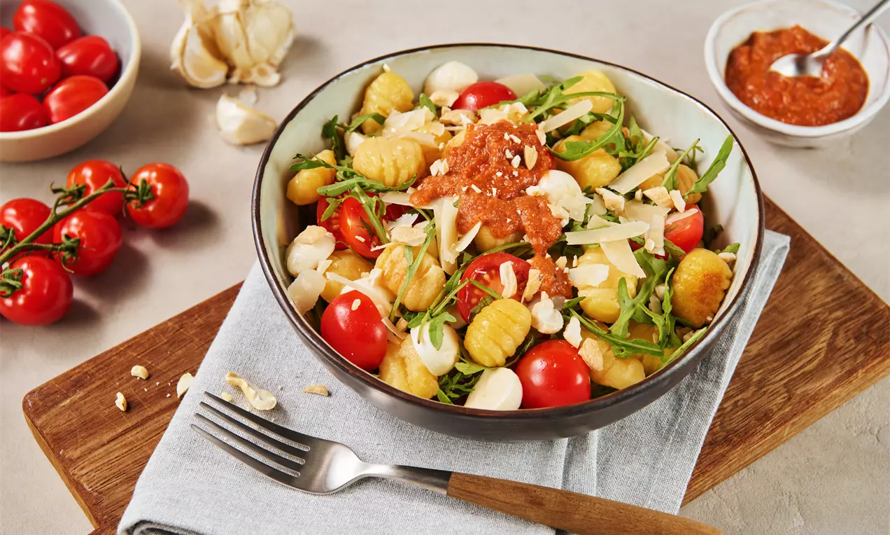 Gnocchi mit goldgelber Kruste, roten Tomaten, Mozzarella, Rucola und cremigem Dip in einer Schüssel auf Holzbrett mit Besteck und Deko