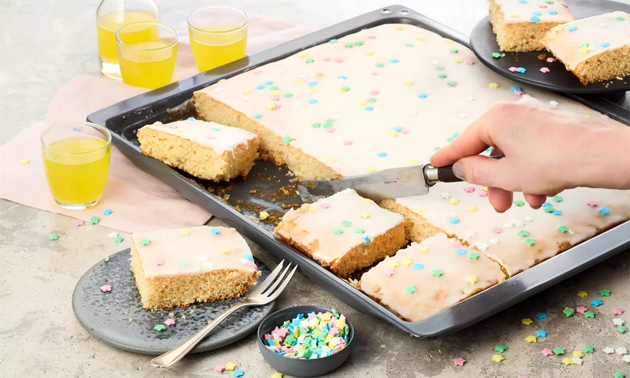 Heller Rührteig mit Zuckerguss und bunten Streuseln auf dem Backblech. Einige Stücke geschnitten und auf einem Teller