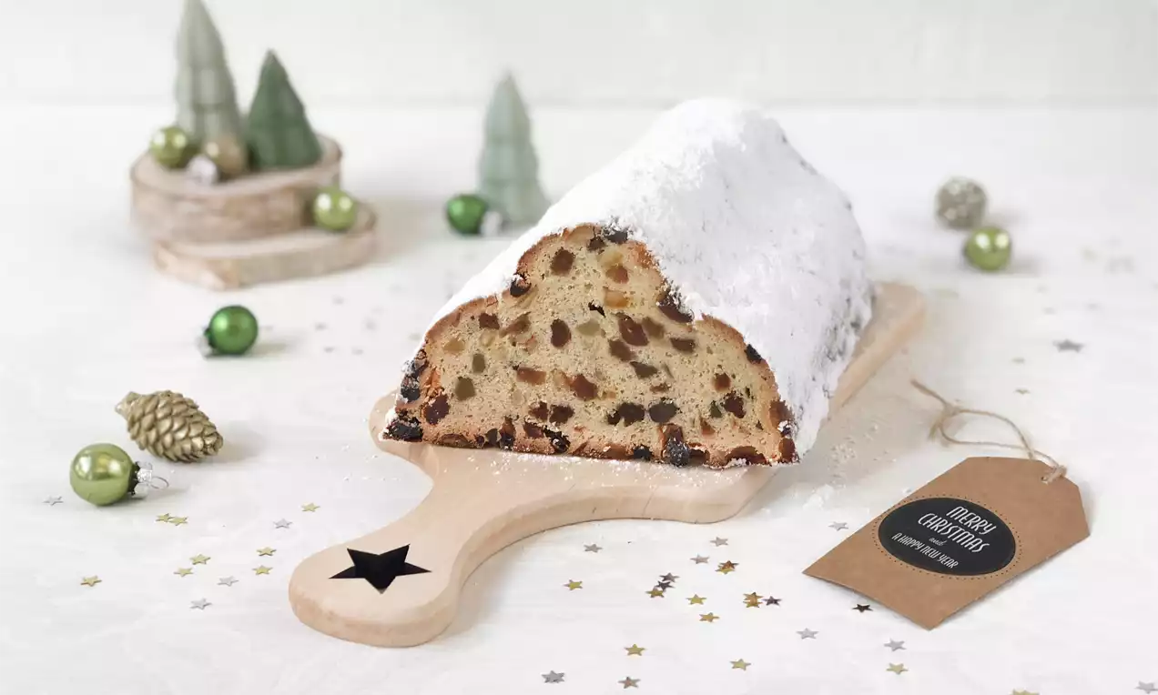 Weihnachtsstollen mit weißem Puderzucker bestäubt auf einem hellen Holzbrett, umgeben von grünen Kugeln und kleinen Tannenbaum-Dekorationen