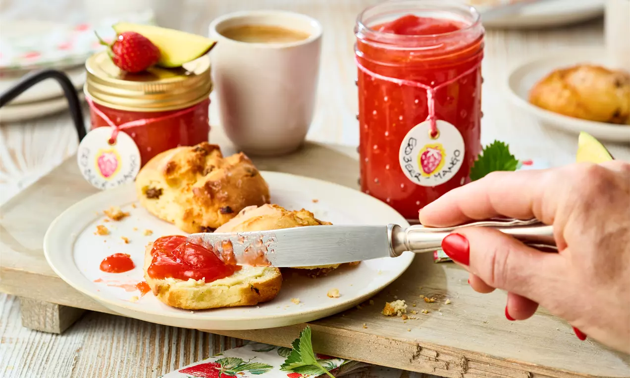 Erdbeer-Mango-Marmelade in einem Glas mit roter Fruchtfüllung und goldfarbenem Deckel auf einem Holzbrett neben Brötchen und Kaffeetasse