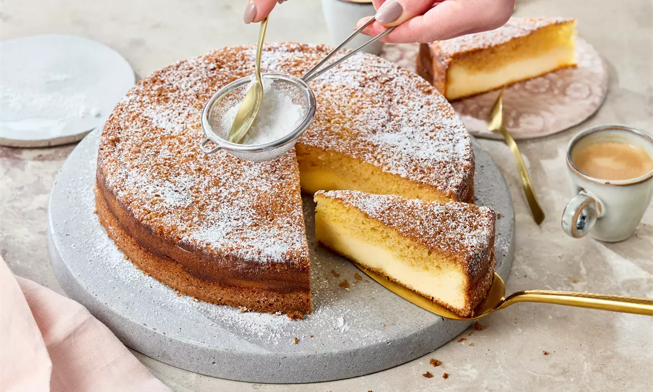 Goldbrauner Dreh-Dich-Um-Kuchen mit heller Füllung, puderzuckerbestäubt, auf einer grauen Platte, daneben Kaffee und Besteck