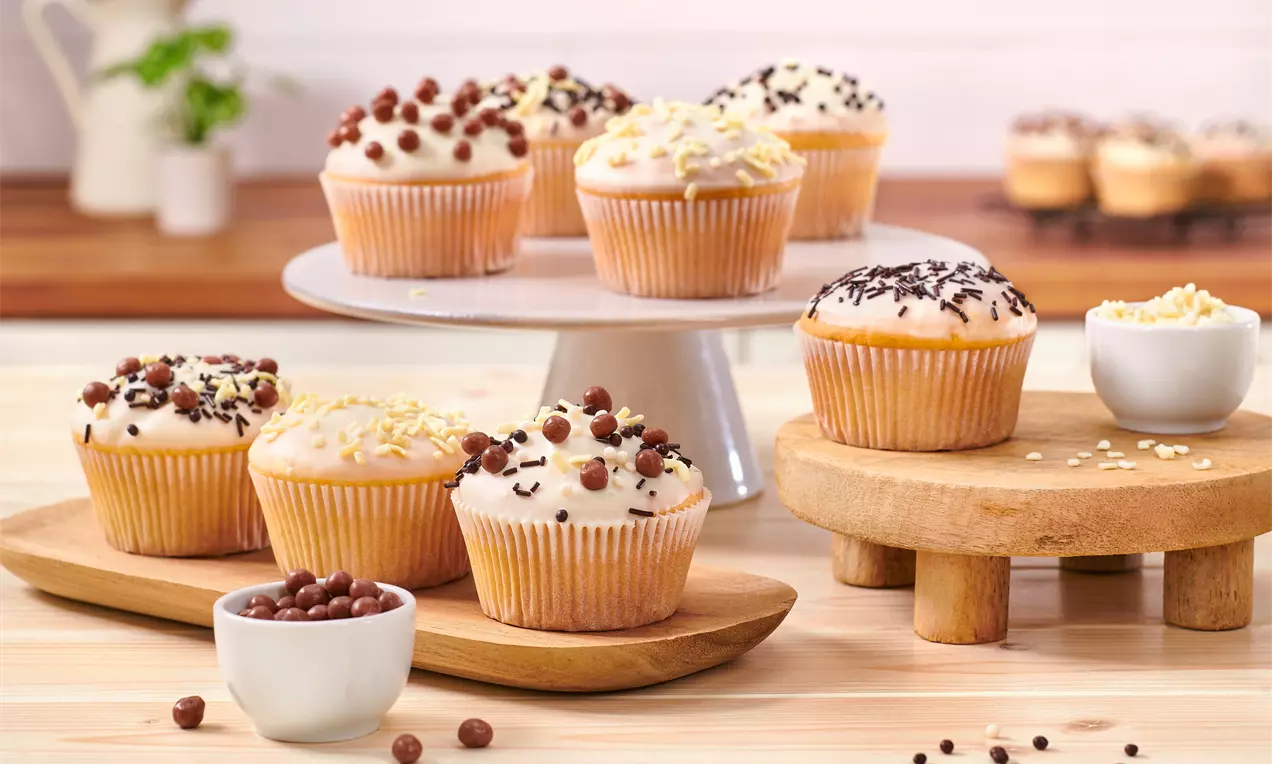 Muffins mit weißer Glasur, dekoriert mit dunklen Schokostreuseln und crispigem Topping, auf Holzplatten und einer weißen Tortenplatte angerichtet