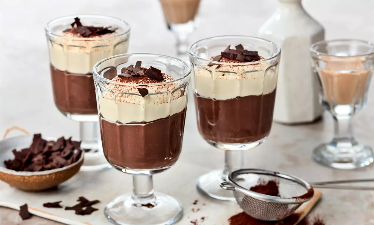 Schokoladiger Pudding in transparenten Gläsern, mit heller Creme bedeckt und dunklen Schokospänen dekoriert auf hellem Untergrund arrangiert
