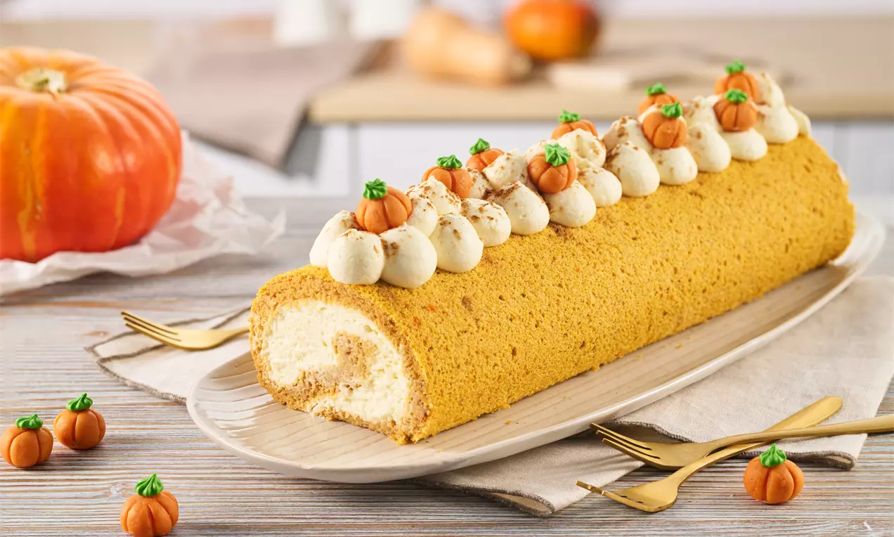 Lockerer, goldbrauner Biskuitkuchen gefüllt mit weißer Creme, dekoriert mit Sahnetupfen, Mini-Kürbissen und Gewürzstaub auf einem Holztablett