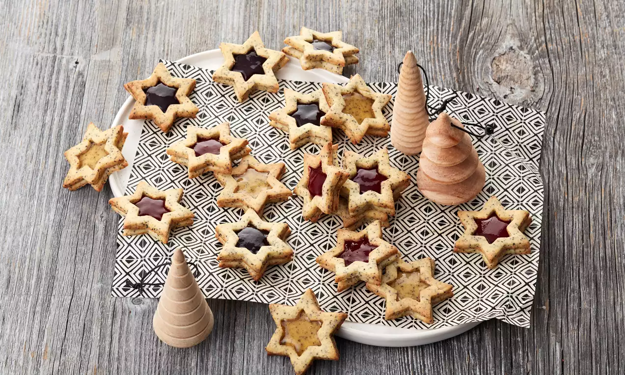 Sterne aus hellem Mohnteig mit fruchtigem, glänzendem Kern auf geometrischem Musterpapier auf weißem Teller, dekoriert mit Holzbäumchen