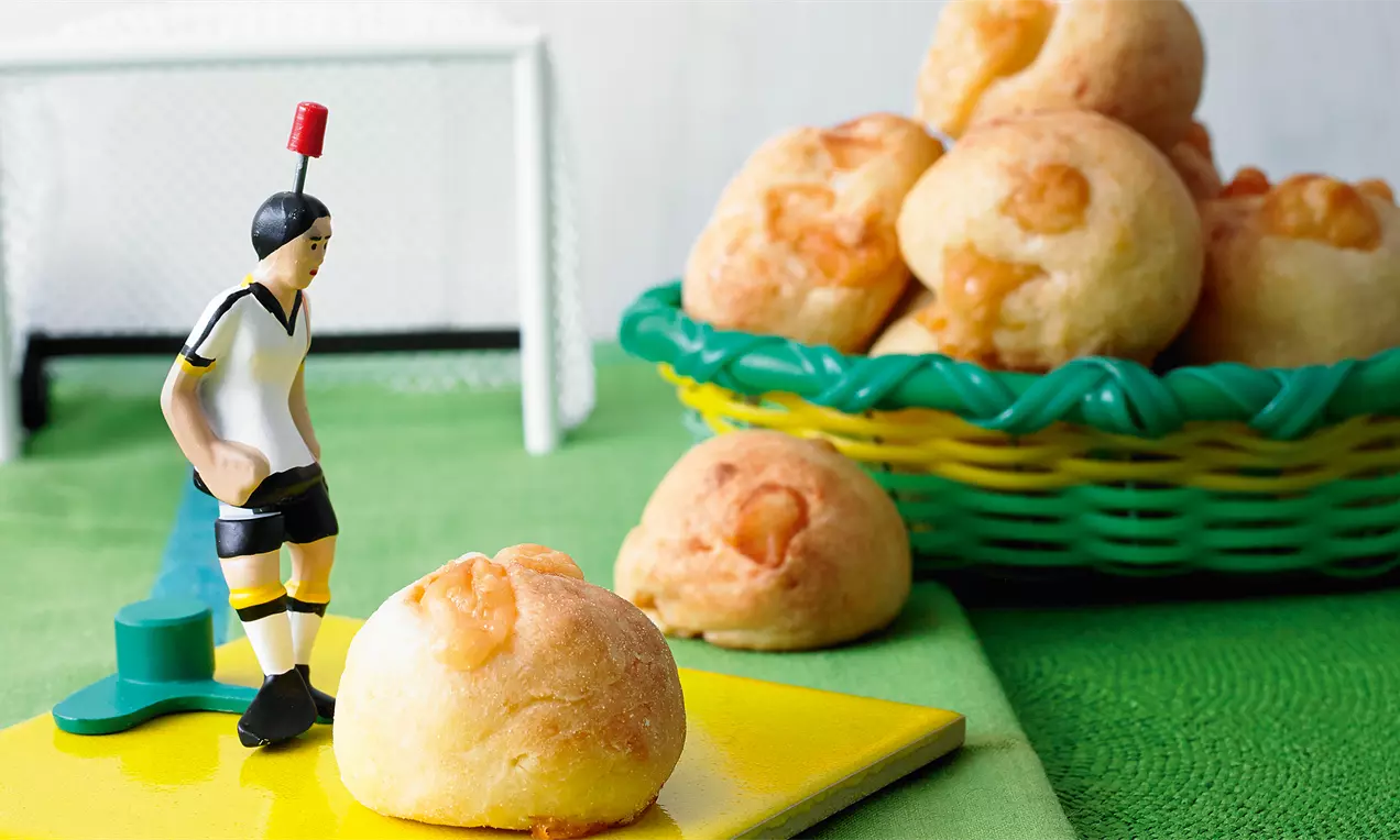 Runde, goldgelbe Käsebrötchen mit leichter Kruste in einem grünen Korb, einige verteilt auf einem gelben Untergrund neben einer Spielfigur